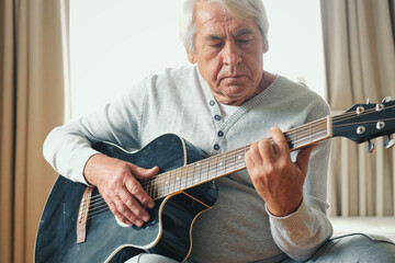 Senior man is playing guitar. Elderly male sitting at sofa and play music. Portrait of a gray...