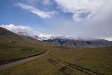 Serene landscape of a dirt road winding through a vast, grassy valley, with snow-capped mountains...