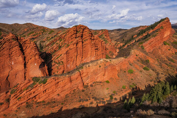 Spectacular red rock formations in the Charyn Canyon, Kazakhstan.  A stunning natural landscape with layered sandstone cliffs and sparse vegetation under a partly cloudy sky.