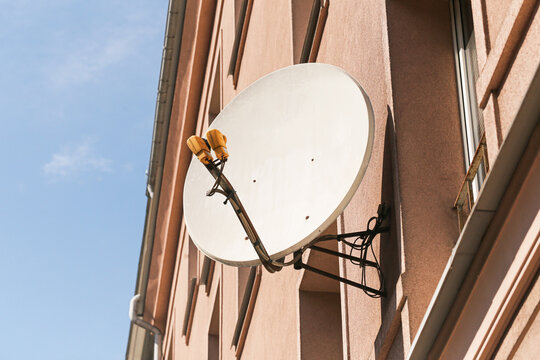 Satellite TV dish on residential building. White receiver antenna against blue sky. Exterior wall mount. Urban apartment communication device. Signal reception equipment for television broadcast.