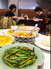 Asian cuisine served in a restaurant, featuring stir-fried morning glory with chili and garlic, spicy tom yum soup, Thai omelet, spring roll, and steamed rice. Three people are sitting
