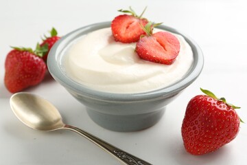 Delicious yogurt in bowl, strawberries and spoon on white table, closeup