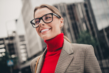 Professional woman in red sweater and plaid blazer smiling outdoors with urban cityscape background