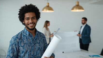 Happy young male architect smiling and holding rolled blueprints in a bright collaborative office setting.
