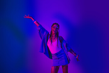 Energetic young woman in stylish denim outfit joyfully posing with vibrant neon lighting in a...