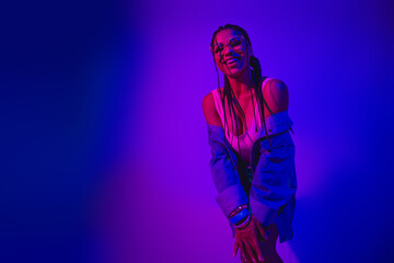 Young fashionable woman enjoying neon lights wearing denim jacket against vibrant gradient purple...