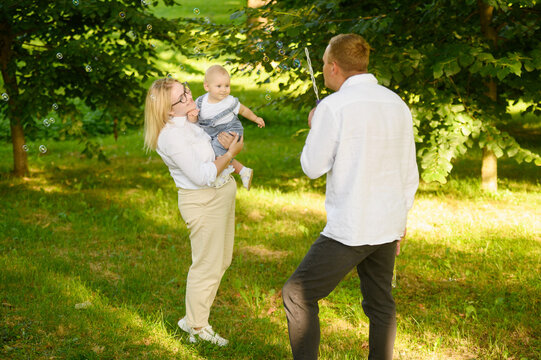 Happy family is relaxing in park. Father is happily playing and blowing soap bubbles, mother is holding cute baby in arms, laughing with him, catching bubbles, playing, enjoying sunny day in nature. - Powered by Adobe