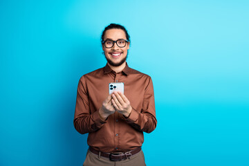Fototapeta premium Young professional man in casual brown formalwear smiling, holding a smartphone against a teal background