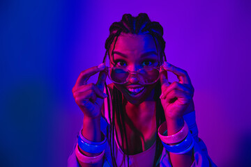 Stylish young woman wearing denim jacket and glasses, illuminated by neon purple and blue lighting...