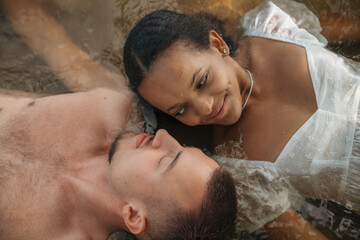 Romantic interracial couple lying in water, heads touching, serene and intimate moment, love,...