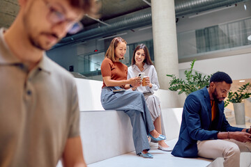 Two businesswomen sitting in office lobby, using smartphone and drinking coffee, while other colleagues are using their phones nearby