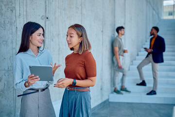 Two businesswomen discussing work while using a tablet in a modern office building, with colleagues in the background