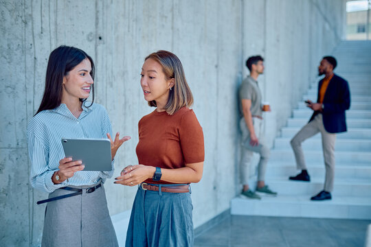 Two businesswomen discussing work while using a tablet in a modern office building, with colleagues in the background