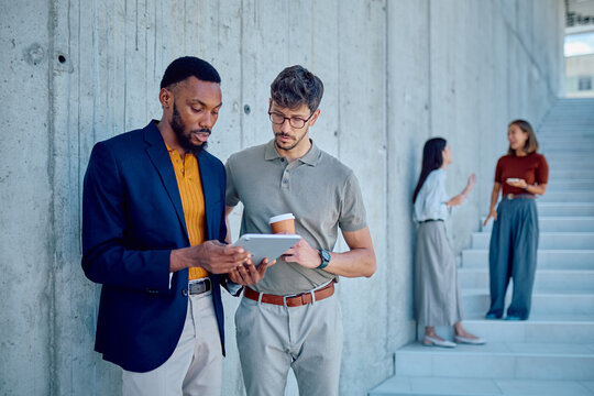 Two businessmen using digital tablet and discussing project in office corridor while their female colleagues talking in background