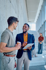 Two smiling businessmen are discussing work while using a digital tablet and drinking coffee in a...