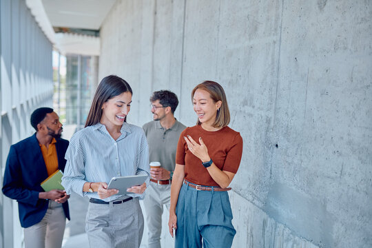 Two businesswomen are walking and talking in a modern office hallway, holding tablet and smiling