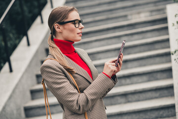 Successful young businesswoman in stylish attire using a smartphone outdoors on urban stairs,...