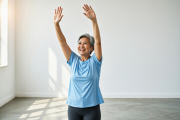 A smiling senior woman with gray hair stretches her arms upwards in a bright, minimalist room.