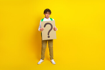 Young schoolboy holding a question mark sign, standing on a vibrant yellow background, symbolizing...