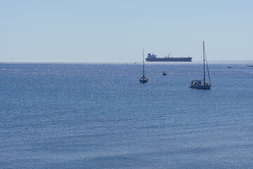 Sailboats gliding across sunlit blue waters, distant tanker hovering near peaceful maritime horizon