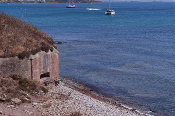 Weathered coastal bunker perched on rocky shoreline, overlooking peaceful bay with sailboats gliding across calm waters