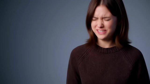A young adult female in a brown sweater displays extreme discomfort or suffering, with a contorted face, closed eyes, and open mouth, standing before a neutral gray backdrop.