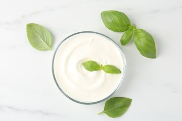 Tasty yoghurt with basil in bowl on white marble table, flat lay