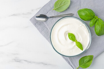 Tasty yoghurt with basil in bowl on white marble table, flat lay. Space for text