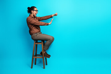 Young man in brown shirt making dynamic gestures while seated on stool with teal background