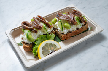Toasted bread topped with anchovies, creamy mozzarella burrata, edamame, and zucchini, finished with lemon zest, isolated on a white marble background. Natural light enhances this healthy seafood-vege