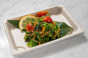 Fresh Japanese wakame seaweed salad in a takeout container on a white marble table with veins. Natural light enhances this healthy Asian dish, perfect for cuisine enthusiasts.


