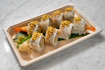 Crunchy Japanese sushi with tempura shrimp, avocado, teriyaki, and pistachio, served in a takeout container on a white marble table with veins. Natural light highlights this fusion delight.

