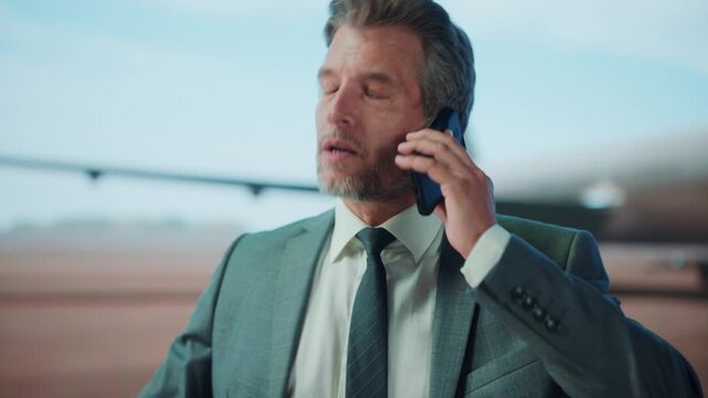 A businessman in a suit talks on a smartphone near an airplane, possibly at an airport, during the day.