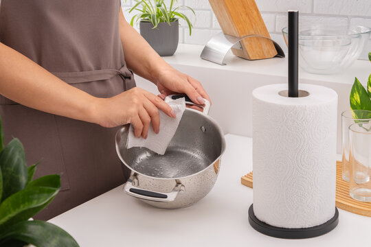 A woman in an apron wipes a shiny metal pot dry with a paper towel. There is a roll, glasses, and plants on the table. Horizontal photo