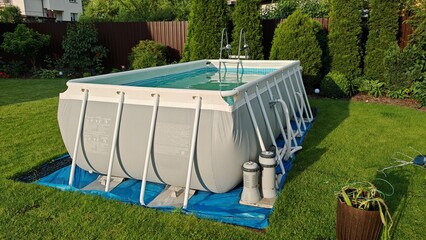 A gray rectangular PVC frame pool stands on a grassy lawn. Nearby is a filter and heater to clean and heat the water. Thuja trees, ornamental bushes and flowers grow next to the metal fence. Sunny