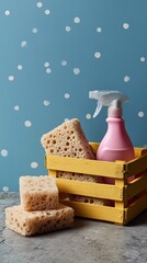 Pink cleaning spray and sponges in a yellow crate on a textured surface with a blue polka dot background.