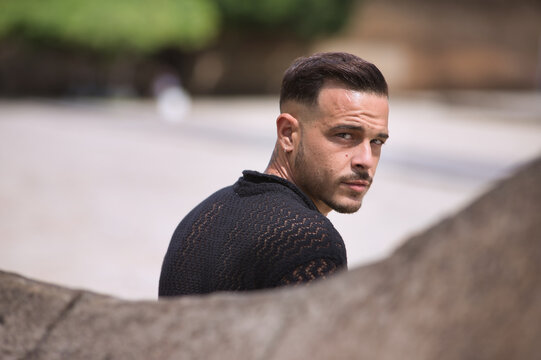 Portrait of young, brunette, Hispanic, modern man in black crochet shirt, with serious and defiant look, his back leaning against a stone wall. Trendy, fashionable, casual concept.