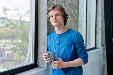 Teenage serious guy with cup in hands in home near window, looking at camera