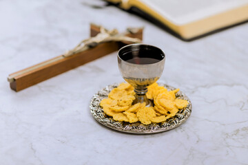 Chalice filled with wine plate of wafers are arranged on marble table with cross an open Bible book.