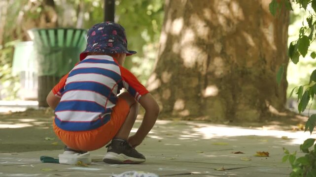 A young boy draws with chalk on a sunlit sidewalk, fully absorbed in his creative play. He is surrounded by lush greenery and large trees in a peaceful park in Simferopol, Crimea.