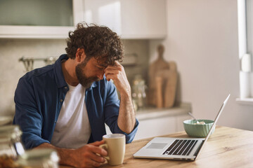A man sits at a wooden kitchen table, holding a coffee cup while looking at a laptop screen with a...