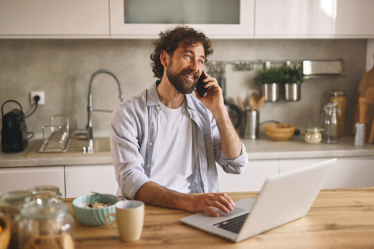 A man with curly hair smiles as he talks on his phone, seated at a kitchen table with a laptop. Morning light shines in, highlighting bowls and a coffee cup nearby. - Powered by Adobe