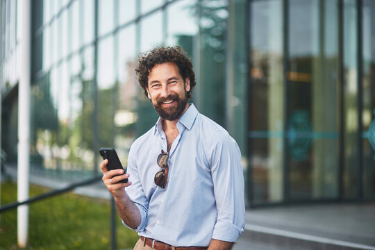 A man in a light blue shirt and sunglasses stands outside a sleek office building, smiling as he uses his smartphone in the warm afternoon light. - Powered by Adobe