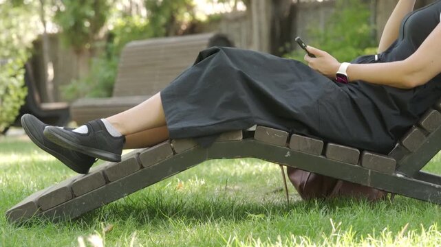 A woman reclines on a wooden lounge chair, using her phone while surrounded by green grass and trees. The peaceful park environment in Simferopol, Crimea, offers a perfect spot for rest and relaxation