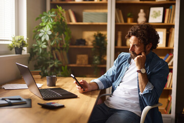 A man is focused on his smartphone while seated at a wooden desk in a home office. Natural light...