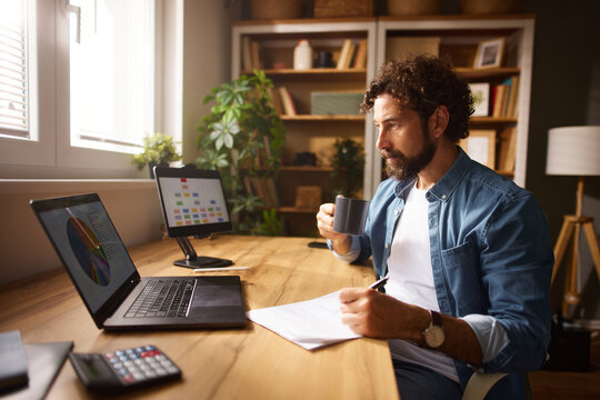 A man calmly engages in work at a neatly organized home office. He holds a coffee cup in one hand while taking notes on a notepad. Laptops display graphs, enhancing his focus on tasks.