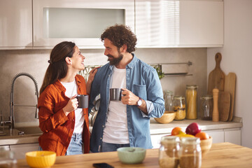 Two people stand close together in a bright home office, each holding a coffee mug and exchanging...