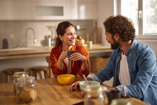A man and woman share a joyful conversation at a wooden table in a stylish home office kitchen, sipping coffee and enjoying a relaxed morning atmosphere.