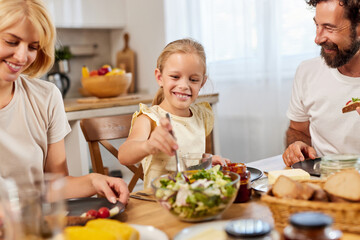 A happy family gathers around the dining table, sharing a meal filled with fresh salad, bread, and...