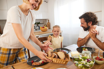 A cheerful family enjoys breakfast in a well-lit kitchen. A woman serves bread while a little girl...